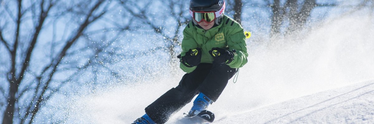 Ein Kind in Skiausrüstung mit Brille und Helm bei der dynamischen Abfahrt auf einem Hügel vor blauem Himmel und winterlichen Bäumen. Der Schnee stäubt durch die Bewegung des Kinds.