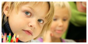 Symbolbild: Ein kleiner blonder Junge mit kurzen Haaren schaut in Nahaufnahme in die Kamera. Vorne sind Buntstifte im Bild. Im Hintergrund sieht man verschwommen ein blondes Mädchen.