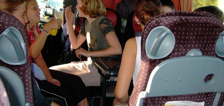 Foto von mehreren jugendlichen in sommerlicher Kleidung. Sie sitzen in einem fahrenden Reisebus und unterhalten sich angeregt.