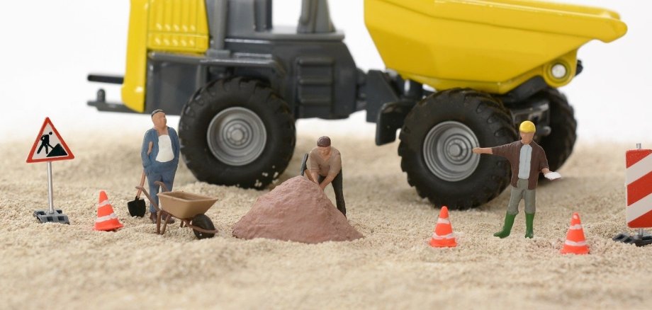 Symbolbild: Foto von Modellbaufiguren als Bauarbeiter, die auf sandigem Untergrund vor einem großen gelben Bagger stehen.