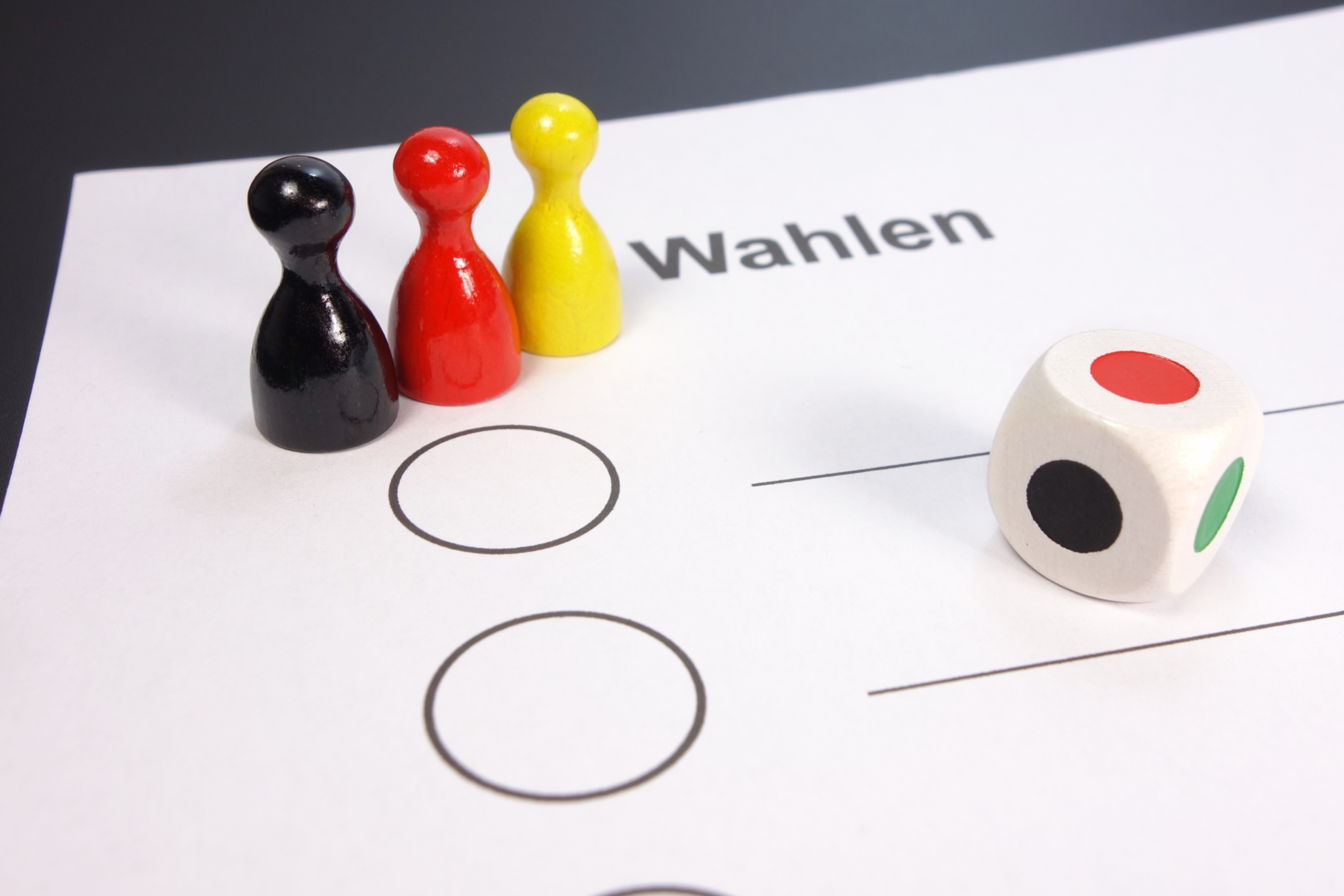 Symbolbild: Foto der oberen linken Ecke eines Wahlscheins mit drei hölzernen Spielfiguren in schwarz, rot und gelb darauf gestellt. Weiter rechts ein weißer Spielwürfel mit Farben statt Augen.