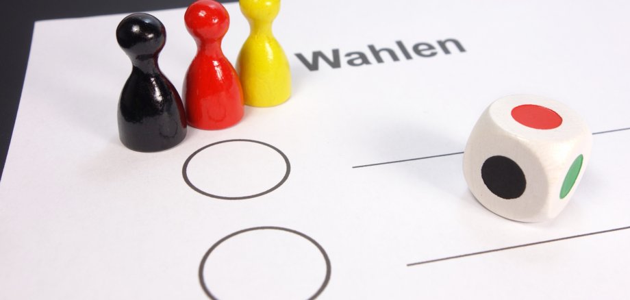 Symbolbild: Foto der oberen linken Ecke eines Wahlscheins mit drei hölzernen Spielfiguren in schwarz, rot und gelb darauf gestellt. Weiter rechts ein weißer Spielwürfel mit Farben statt Augen.