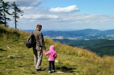 Foto: Rückansicht eines Mannes in moderner Wanderkleidung, er hält ein Kind in rosa Jacke an der Hand. Sie stehen auf einer abschüssigen Bergwiese im Sonnenschein und schauen auf das Harzpanorama.